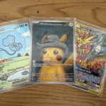 Drei Pokémon-Sammelkarten mit Pikachu, Mew und den Legendären Vögeln in Hüllen auf Holztisch.
