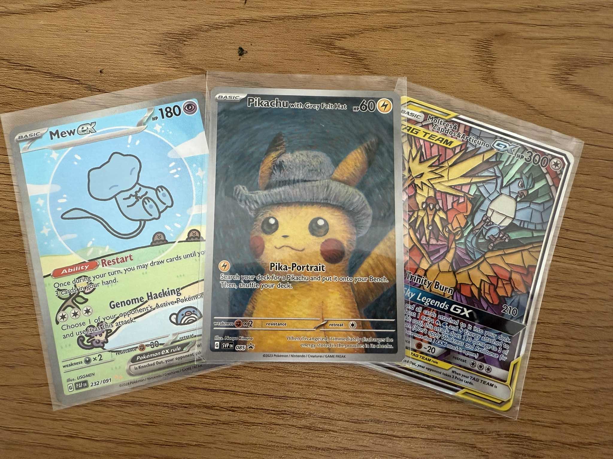 Drei Pokémon-Sammelkarten mit Pikachu, Mew und den Legendären Vögeln in Hüllen auf Holztisch.
