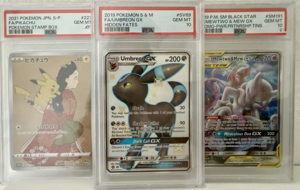 Drei Pokémon-Sammelkarten mit PSA-10-Bewertung in Acrylhüllen: Pikachu, Umbreon GX und Mewtwo & Mew GX.