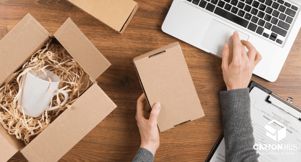 Person verpackt eine weiße Tasse in Karton mit Füllmaterial am Schreibtisch neben Laptop